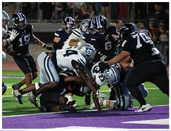 A person in a blue football uniform and white helmet with an American flag design is crouched in a ready position on a football field. Descripci n generada con IA
