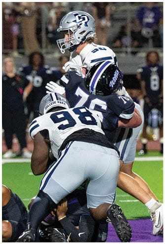 A person in a white and blue football uniform is lunging forward while being blocked by a person in a dark blue uniform during a football game on a green field. Descripci n generada con IA