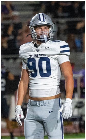 A person wearing a football helmet and navy blue jersey with “COLLEGE PARK" and the number 8 is on a field with blurred figures in the background. Descripci n generada con IA
