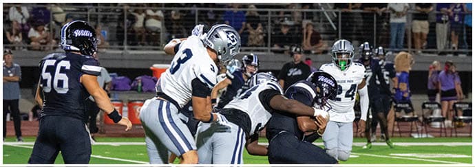 People wearing football uniforms are engaged in a play on a football field with one person in dark clothing being blocked by others in white and blue uniforms. Descripci n generada con IA