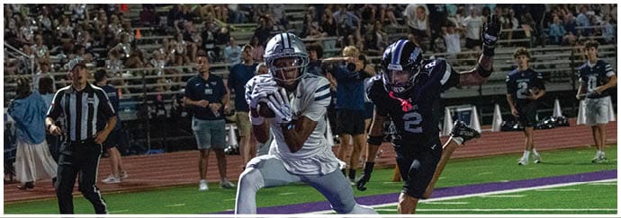 People wearing football uniforms are engaged in a play on a football field with one person in dark clothing being blocked by others in white and blue uniforms. Descripci n generada con IA