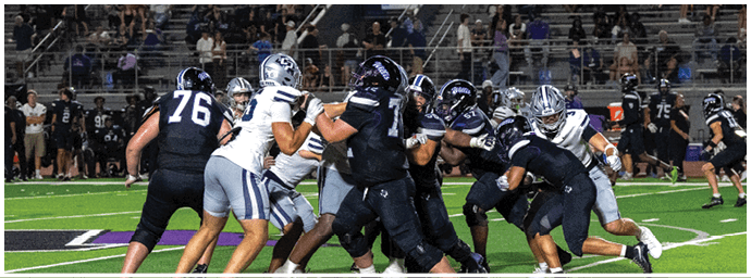 People wearing helmets and football uniforms are engaged in a play on a green football field with bleachers and spectators in the background. Descripci n generada con IA