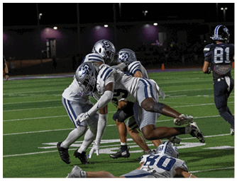 A person wearing a white football helmet and a navy blue jersey with “COLLEGE PARK" written on it is standing on a field with other players in the background. Descripci n generada con IA