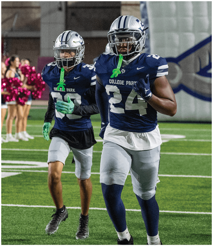 Two people wearing dark blue football uniforms with helmets and faceguards stand on a field, one prominently displaying the number 24 and the words “College Park" on the jersey. Descripci n generada con IA