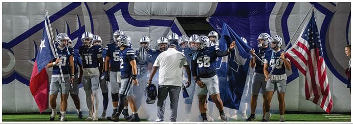A group of people in football uniforms and helmets stand on a field holding flags, accompanied by a person in casual clothing walking among them. Descripci n generada con IA