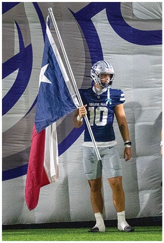 A person in a football uniform and helmet stands on a field holding a large Texas state flag. Descripci n generada con IA
