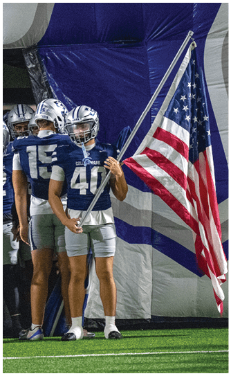 People in football uniforms stand together with one holding an American flag inside a sports facility. Descripci n generada con IA