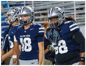 Two people wearing blue College Park football uniforms and helmets stand on a sports field with bleachers in the background. Descripci n generada con IA