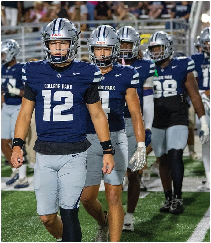 Four people wearing blue football uniforms and silver helmets stand in a row on a field with blurred spectators in the background. Descripci n generada con IA