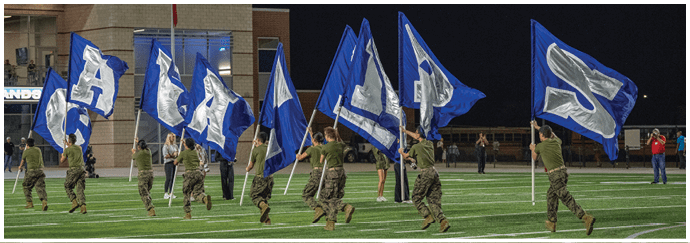 People in military uniforms are running on a sports field at night while holding large blue flags with white letters. Descripci n generada con IA