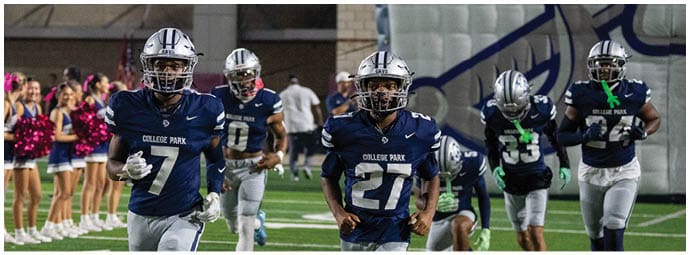 Five people in football uniforms with helmets are running on a field with cheerleaders in the background. Descripci n generada con IA