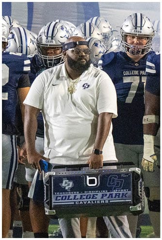 A person wearing a white polo shirt and visor stands holding a large case with a College Park Cavaliers logo while surrounded by people in football uniforms on a field. Descripci n generada con IA