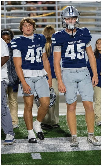 Two people wearing College Park football uniforms stand side by side on a field, one holding a helmet and the other wearing a helmet. Descripci n generada con IA