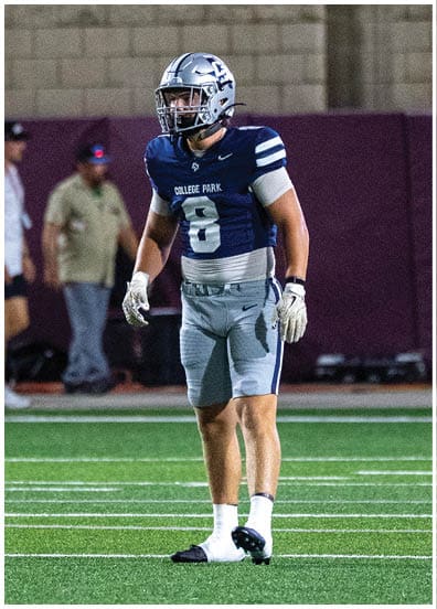 A person wearing a football uniform with a helmet and gloves stands on a green field with white yard lines during a game. Descripci n generada con IA