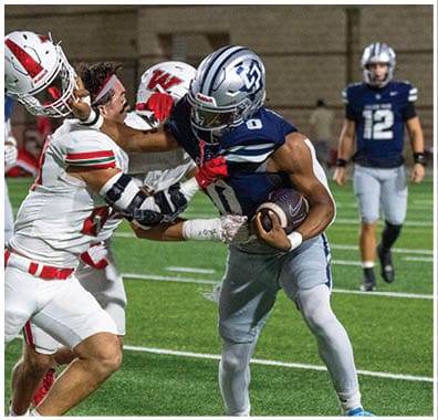 Two people in football uniforms are engaged in a tackle on a field while other players watch in the background. Descripci n generada con IA