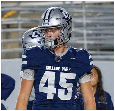 A person wearing a silver football helmet and a navy blue jersey with white stripes is looking to the side on a sports field. Descripci n generada con IA