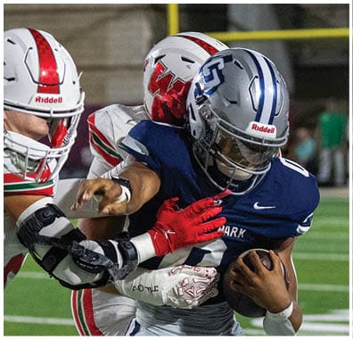 Three people wearing football helmets and uniforms are engaged in a play on a football field, with one person in a blue uniform holding the ball while two others in white uniforms attempt to tackle them. Descripci n generada con IA