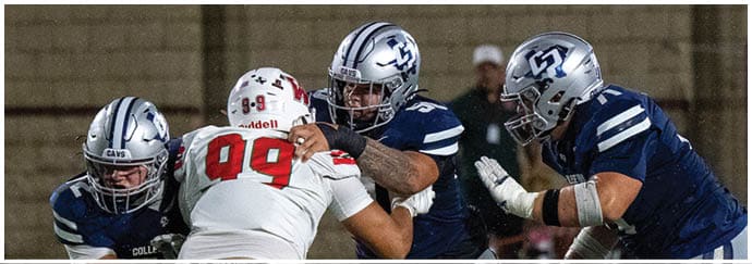 People in football uniforms are engaged in a play on a field with one person in white attempting to block or tackle players in blue. Descripci n generada con IA