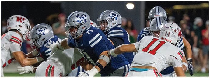 People in football uniforms are engaged in a play on a field with some players blocking and others preparing to move. Descripci n generada con IA