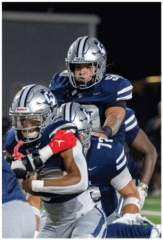 Three football players in blue and silver uniforms are engaged in a play during a nighttime game, with one player holding the ball and others positioned closely around. Descripci n generada con IA