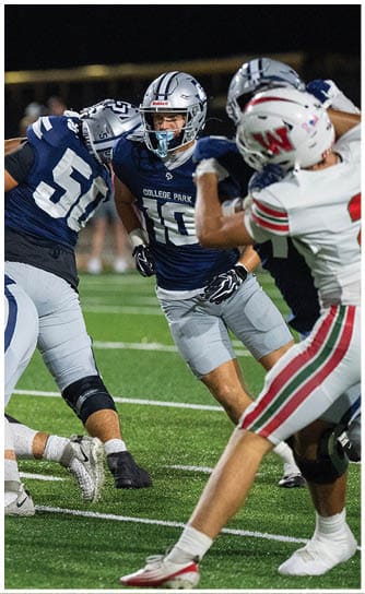 A person wearing a blue College Park football uniform and helmet is actively engaged in a play against opponents in white and red football uniforms on a field. Descripci n generada con IA