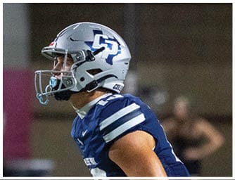 A person wearing a blue and white football uniform and helmet is focused and appears to be preparing for action on a football field. Descripci n generada con IA