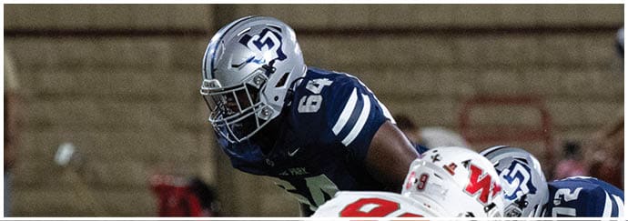 A person wearing a blue football uniform and a silver helmet with a logo is leaning forward on a field with a blurred background. Descripci n generada con IA