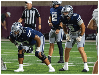 Two people in blue and gray football uniforms are positioned on a field ready for a play with a referee and another player in the background. Descripci n generada con IA