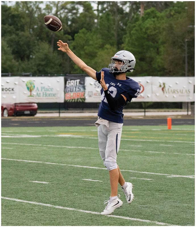 A person wearing a helmet and sports uniform is throwing a football on a grassy field with trees and banners in the background. Descripci n generada con IA