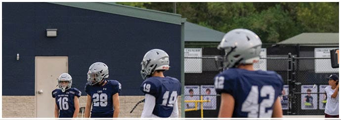 Five people wearing blue football uniforms and helmets stand on a field near an orange yard marker with a building and trees in the background. Descripci n generada con IA