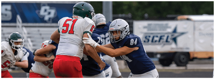 People wearing football uniforms are engaged in a play on a football field with one person in a blue jersey blocking another in a white and red jersey. Descripci n generada con IA