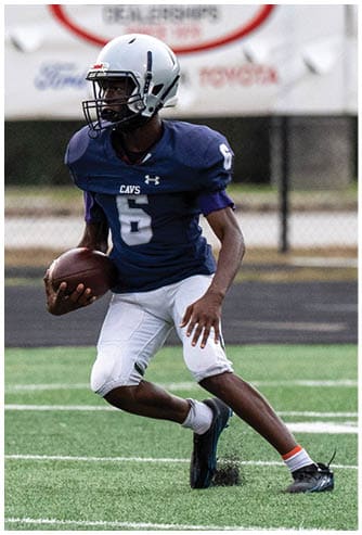 A person wearing a blue football jersey with the number six and a white helmet is running on a football field while holding a football. Descripci n generada con IA