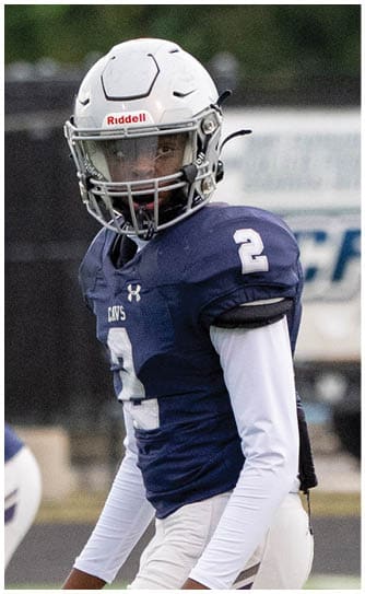 A person wearing a white football helmet and a navy blue jersey with the number 2 is looking towards the camera on a sports field. Descripci n generada con IA