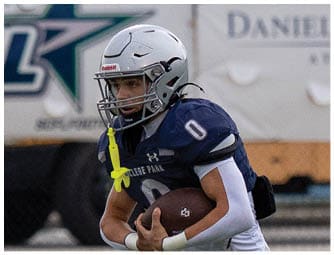 A person wearing a white football helmet and navy blue jersey is on a field with a blurred sports advertisement banner in the background. Descripci n generada con IA