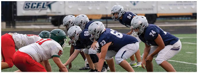 People in football uniforms from two opposing teams are lined up facing each other on a field, preparing for a play. Descripci n generada con IA