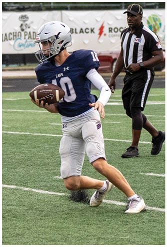 A person wearing a blue football jersey and white helmet is running on a football field while holding a football, with a referee walking behind them. Descripci n generada con IA