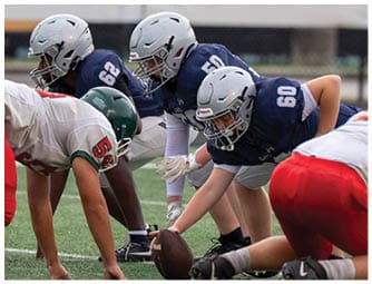 Adults in football uniforms are lined up on a field preparing for a play with one player holding the football ready to snap it. Descripci n generada con IA