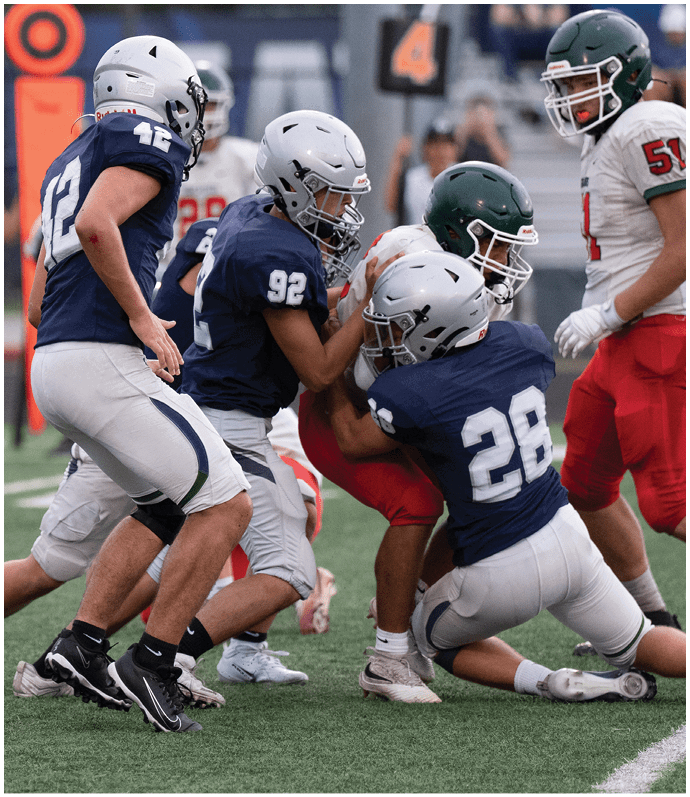 Several people wearing football uniforms are engaged in a tackle on a grassy field during a game. Descripci n generada con IA