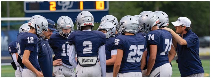 A group of football players in blue and white uniforms huddle together on a field while a coach in a blue shirt and white cap speaks to them. Descripci n generada con IA