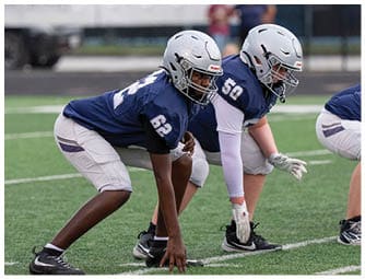 Three people in football uniforms are crouched in a ready position on a grassy field, preparing for a play. Descripci n generada con IA