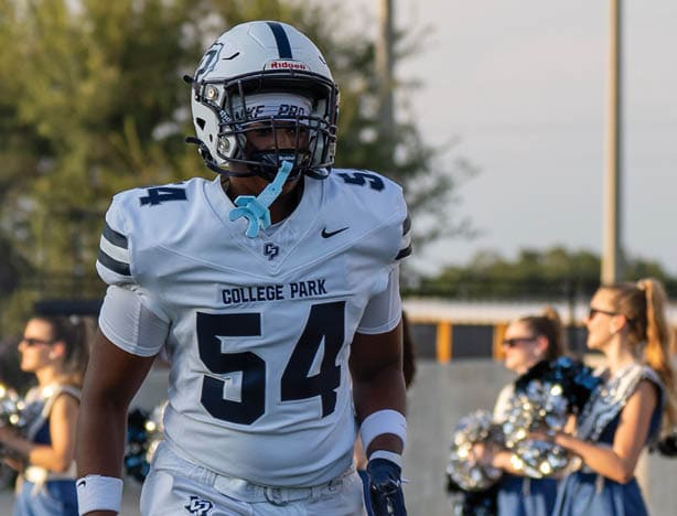 A person in a white football uniform with the number 54 and a helmet is walking on a field with cheerleaders in blue and white uniforms holding pom-poms in the background. Descripci n generada con IA