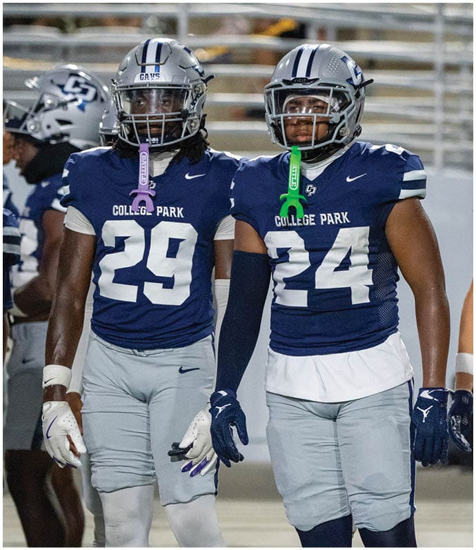 Two football players in blue and gray College Park uniforms stand side by side on a field with helmets and mouthguards. Descripci n generada con IA