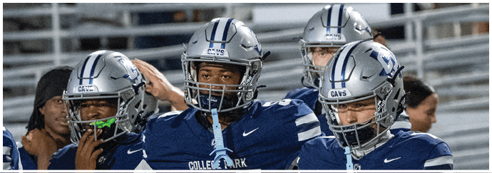 Three people wearing blue football uniforms and helmets stand together on a sports field with bleachers in the background. Descripci n generada con IA