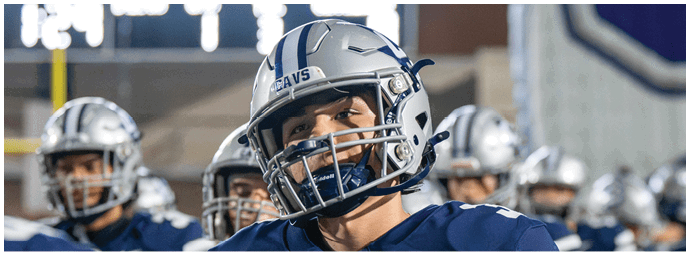 A group of people wearing football helmets and navy blue jerseys stand together on a field, with one person in the foreground smiling. Descripci n generada con IA