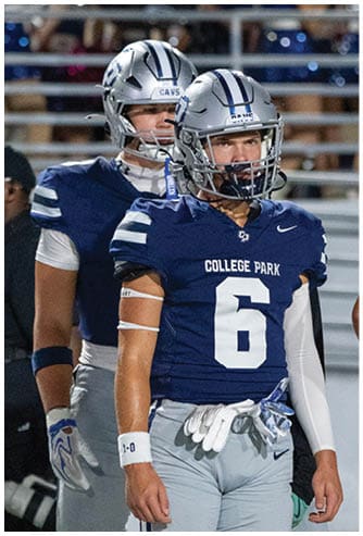 Two people in football uniforms and helmets stand on a field with a person in black clothing and a cap in the background. Descripci n generada con IA