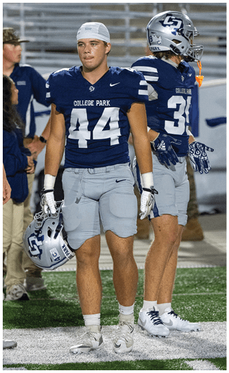 A person wearing a College Park football uniform with the number 44 stands on a field holding a helmet, surrounded by other players and staff. Descripci n generada con IA