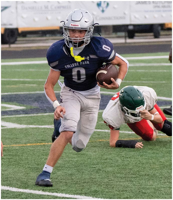 A person in a blue and white football uniform is running with the ball while two people in white and red football uniforms are attempting to block or tackle on a football field. Descripci n generada con IA