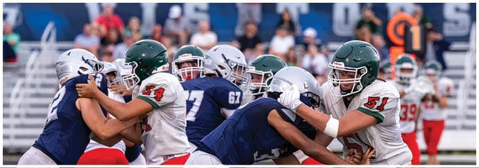 People wearing football helmets and uniforms are engaged in a tackle during a game on a field with bleachers and spectators in the background. Descripci n generada con IA
