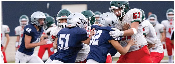 People wearing helmets and football uniforms are engaged in a close physical play on a football field with a referee and other players in the background. Descripci n generada con IA