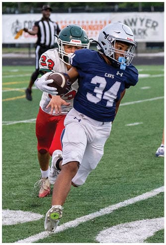 A person in a blue football uniform with the number 34 is running with the ball while being pursued by another person in a red and white uniform on a football field. Descripci n generada con IA